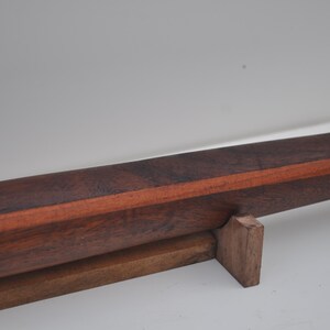 May include: A handcrafted wooden object with a dark brown exterior and a reddish-brown stripe. The object is supported by a smaller, lighter brown wooden base. The wood grain is visible, suggesting a natural finish.