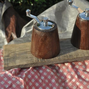 May include: Two wooden pepper grinders with silver-colored tops and handles sit on a weathered wooden board. The grinders are a rich brown color and have a classic, conical shape. The background includes a red and white checkered cloth.