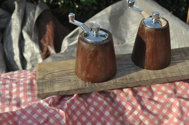 May include: Two wooden pepper grinders with silver-colored tops and handles sit on a weathered wooden board. The grinders are a rich brown color and have a classic, conical shape. The background includes a red and white checkered cloth.