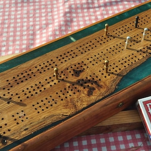 May include: A wooden cribbage board with a green and brown epoxy resin inlay. The board has brass and black pegs and is ready for a game. A deck of Bicycle playing cards is in the background.