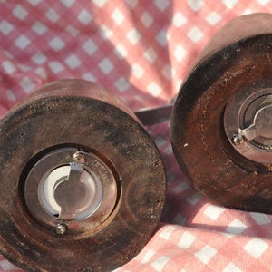 May include: Close-up of two wooden salt and pepper grinders. The grinders are cylindrical with a dark brown finish and metal grinding mechanisms. The image is set against a red and white checkered background.