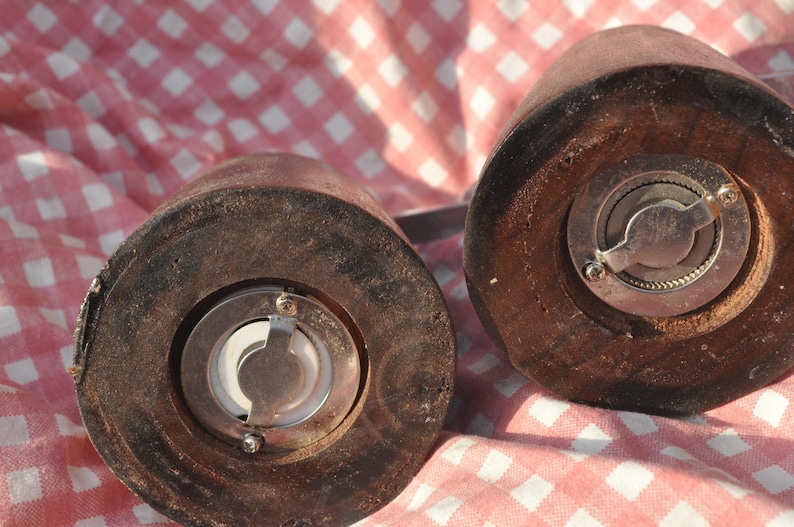 May include: Close-up of two wooden salt and pepper grinders. The grinders are cylindrical with a dark brown finish and metal grinding mechanisms. The image is set against a red and white checkered background.