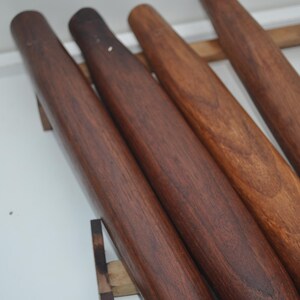 May include: Four wooden rolling pins of varying shades of brown are displayed. The rolling pins are cylindrical with rounded ends. The wood grain is visible, and the pins are arranged on a wooden rack against a white background.