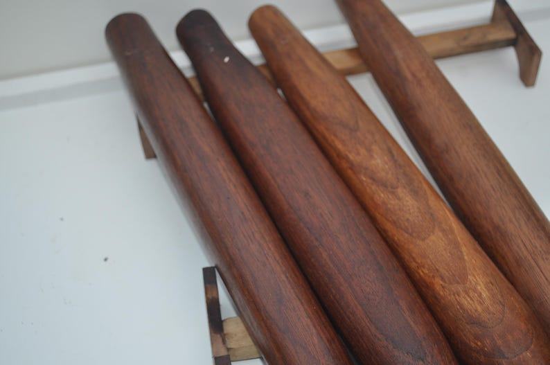 May include: Four wooden rolling pins of varying shades of brown are displayed. The rolling pins are cylindrical with rounded ends. The wood grain is visible, and the pins are arranged on a wooden rack against a white background.