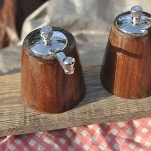May include: Two wooden salt and pepper grinders with stainless steel tops and crank handles. The grinders are made of dark brown wood and sit on a wooden plank. The background includes a red and white checkered cloth.