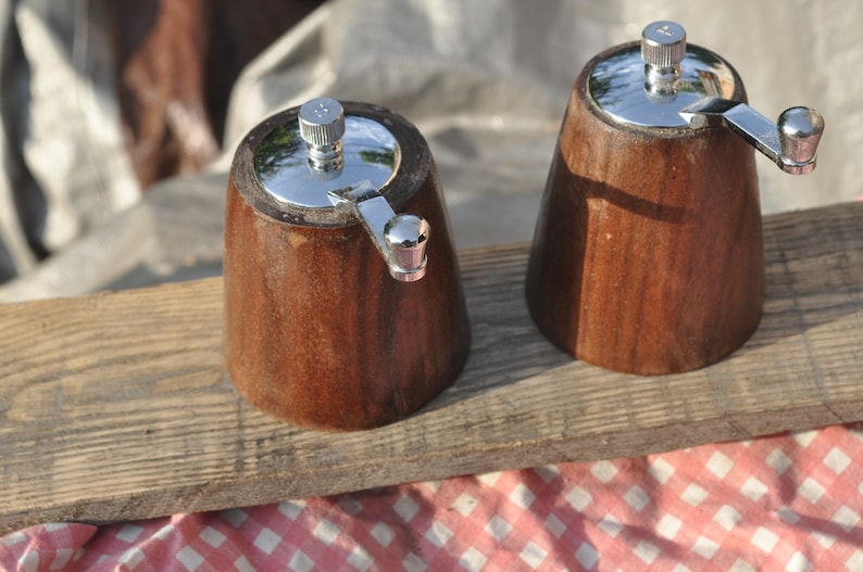 May include: Two wooden salt and pepper grinders with stainless steel tops and crank handles. The grinders are made of dark brown wood and sit on a wooden plank. The background includes a red and white checkered cloth.