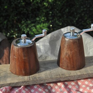 May include: Two wooden salt and pepper grinders with stainless steel tops and crank handles. The grinders are a rich brown color and sit on a weathered wooden board. The background is a blurred green.