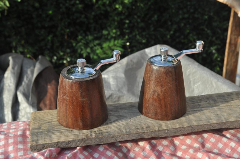 May include: Two wooden salt and pepper grinders with stainless steel tops and crank handles. The grinders are a rich brown color and sit on a weathered wooden board. The background is a blurred green.