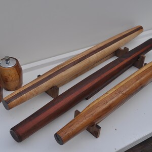May include: Wooden rolling pins in various shades of brown, with a dark stripe down the center of some. Two wooden pepper grinders with silver tops are also present. The rolling pins are supported by small wooden stands.