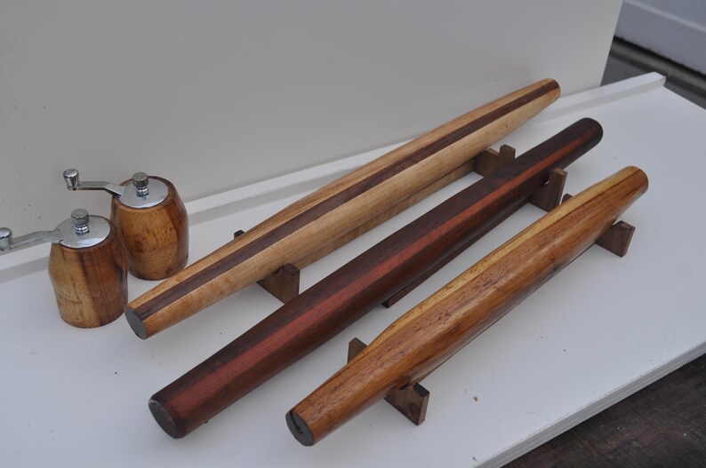 May include: Wooden rolling pins in various shades of brown, with a dark stripe down the center of some. Two wooden pepper grinders with silver tops are also present. The rolling pins are supported by small wooden stands.