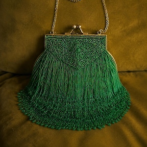 May include: A vibrant emerald green beaded handbag featuring a structured frame with a gold-toned metal clasp closure and a matching delicate chain strap. The body of the bag is covered in intricate beadwork with long, flowing fringe tassels.