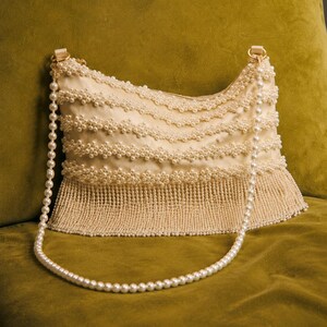 May include: A cream-colored handbag featuring horizontal rows of white faux pearls across the body and a dense fringe of small white beads along the bottom edge. The bag includes a long shoulder strap made of a single strand of white faux pearls.
