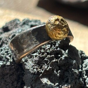 Può includere: Un anello caratterizzato da una larga fascia in metallo scuro martellato con un elemento circolare color oro dalla superficie testurizzata sulla parte superiore. La fascia ha una finitura opaca e vissuta, mentre l'elemento dorato presenta una superficie irregolare.