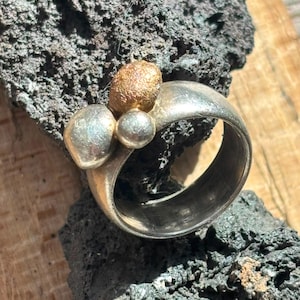 Può includere: Un anello in argento con una fascia larga e tre distinti accenti sferici sulla parte superiore. Una sfera presenta una finitura testurizzata color bronzo, mentre le altre due sfere sono lisce e color argento, creando un aspetto multidimensionale.