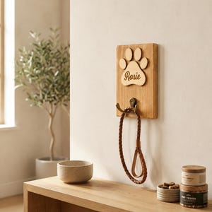 May include: A rectangular wooden wall-mounted hook featuring a light-colored wood grain finish. Attached to the top is a wooden paw print cutout engraved with the name Rosie. Below the paw print is a dark metal hook holding a braided brown leash.