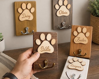 May include: Five rectangular wooden wall hooks, each featuring a light-colored wooden paw print cutout engraved with a pet name: Daisy, Olivia, Buster, Sadie, or Cooper. Each plaque includes a metal double hook for hanging items.