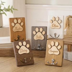 Puede incluir: Cinco ganchos de pared rectangulares de madera con un diseño de huella de pata en relieve. Cada placa está grabada con un nombre: Buster, Sadie, Olivia, Daisy y Cooper. Los soportes tienen varios tonos de madera y blanco, con un gancho metálico.