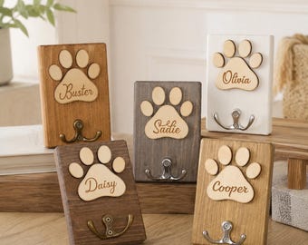 Può includere: Cinque ganci da parete rettangolari in legno con un design a zampa in rilievo. Ogni targa è incisa con un nome: Buster, Sadie, Olivia, Daisy e Cooper. I supporti sono rifiniti in varie tonalità di legno e bianco, ciascuno con un gancio in metallo.