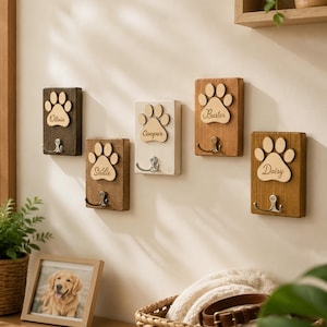 May include: Five rectangular wooden wall hooks, each featuring a light-colored wooden paw print cutout engraved with the names Olivia, Sadie, Cooper, Buster, and Daisy. Each plaque includes a single metal hook for hanging items, with varying wood stains ranging from dark brown to light white-washed finishes.