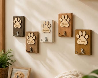 May include: Five rectangular wooden wall hooks, each featuring a light-colored wooden paw print cutout engraved with the names Olivia, Sadie, Cooper, Buster, and Daisy. Each plaque includes a single silver-toned metal hook for hanging items.