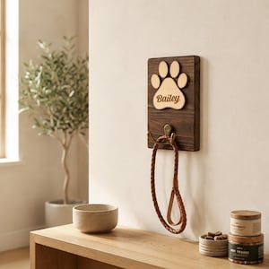 May include: A rectangular dark brown wooden wall plaque featuring a light-colored wooden paw print cutout with the name Bailey engraved in the center. A brass-toned metal hook is attached below the paw print, holding a braided brown leather leash.