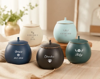 May include: Five round ceramic pet urns with lids in navy blue, cream, light blue, black, and dark green. Each urn features custom engraved text, including names like Bruce, Charlie, and Toby, along with paw print and angel wing motifs.
