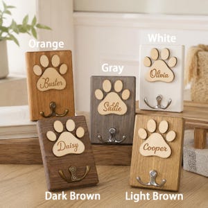 May include: Five rectangular wooden wall hooks, each featuring a light-colored wooden paw print cutout with a name engraved in cursive script. The hooks are finished in orange, gray, white, dark brown, and light brown, each with a metal hook.