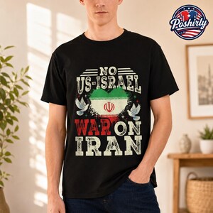 Op de afbeelding: Een zwart t-shirt met een afbeelding en de tekst NO US-ISRAEL WAR ON IRAN. Het ontwerp bevat een hartvormige Iraanse vlag, twee witte duiven en versleten witte en rode letters die in het midden op de borst van het kledingstuk staan.