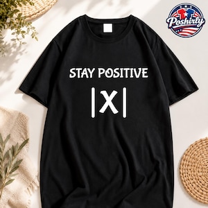May include: A black short-sleeved t-shirt featuring the white text STAY POSITIVE centered on the chest, positioned above a white mathematical absolute value symbol enclosing the letter X, displayed as |X|.