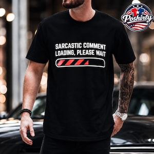 Puede incluir: Una camiseta negra de manga corta con el texto blanco "SARASTIC COMMENT LOADING, PLEASE WAIT" sobre un gráfico de barra de carga con contorno blanco, parcialmente relleno con rayas diagonales rojas desde el lado izquierdo.