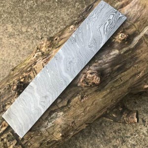 May include: A rectangular metal billet featuring a distinct gray and silver Damascus steel pattern with flowing, wavy lines across the entire surface. The flat piece of metal has sharp, straight edges and a polished finish that highlights the layered steel.
