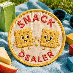 Può includere: Una toppa ricamata circolare con sfondo color crema e bordo giallo. Presenta due biscotti quadrati color marrone dorato, sorridenti, con braccia e gambe, e il testo ricamato in rosso SNACK DEALER arcuato sopra e sotto di essi.