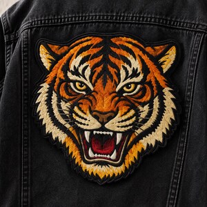 May include: A black denim jacket featuring a large, detailed embroidered patch of a tiger head on the back. The tiger is depicted with orange, black, and cream-coloured thread, showing a snarling expression with visible teeth and yellow eyes.