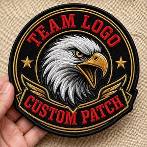 May include: A circular embroidered patch featuring a central eagle head with white feathers and a yellow beak. The black background includes the red text TEAM LOGO at the top and CUSTOM PATCH at the bottom, accented by two gold stars and gold wings.