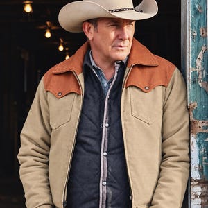 May include: A beige cowboy hat with a dark braided band, a tan jacket featuring rust-colored shoulder yokes and a front zipper, a dark gray quilted vest with silver snap buttons, and a light blue collared shirt worn underneath the layered outerwear.
