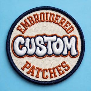 Peut inclure: Un écusson brodé circulaire avec une base en tissu crème et une bordure cousue bleu foncé. Le texte EMBROIDERED est brodé en orange en haut, CUSTOM en grandes lettres blanches avec des contours orange au centre, et PATCHES en orange en bas.