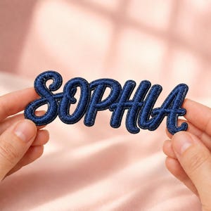 Può includere: Una toppa ricamata blu scuro che riporta il nome SOPHIA in un carattere corsivo collegato. Le lettere hanno un aspetto strutturato e in rilievo con un bordo cucito distinto che delinea ogni lettera, creando un look audace e definito.