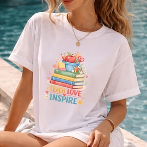 May include: A white short-sleeved t-shirt featuring a colourful graphic of stacked books, a red apple, and school supplies. The text "TEACH LOVE INSPIRE" is printed in bold, multi-coloured letters below the illustration, surrounded by small hearts and flowers.