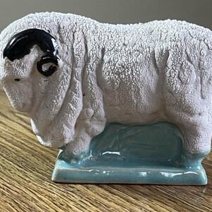 May include: A ceramic sheep figurine featuring a textured white body with a matte finish, a glossy black curved horn, and a smooth light blue base. The sheep is depicted in a standing position, showcasing detailed wool-like surface texture.