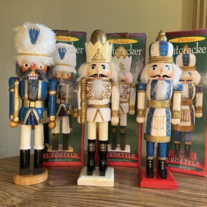 May include: Three decorative nutcracker figures standing in front of their individual boxes. The left figure wears a blue uniform and white fur hat. The center figure is white and gold with a crown. The right figure wears a blue and white uniform.
