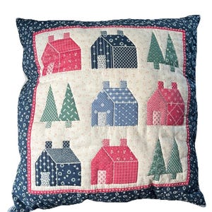 May include: A square throw pillow featuring a patchwork design of houses and evergreen trees in shades of red, navy blue, and green on a cream background. The border is a dark blue fabric with a small white floral pattern and a thin red trim.
