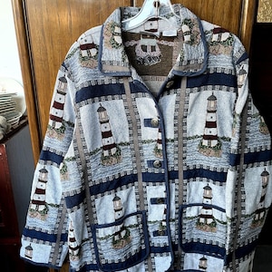 May include: A long-sleeved button-down jacket featuring a woven tapestry pattern of red and white lighthouses on a light blue and navy background. The garment includes a collared neckline, two front patch pockets, and metallic buttons.