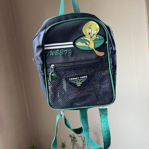 May include: A black mini backpack with green trim and straps, featuring an embroidered Tweety Bird character, the text "TWEETY" in green, and a front mesh pocket with a "LOONEY TUNES" patch. The bag includes a top handle and two adjustable shoulder straps.
