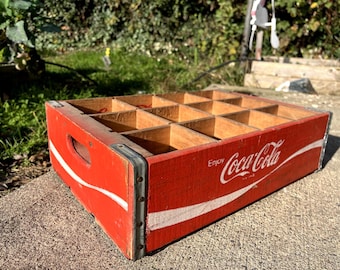 May include: A rectangular wooden Coca-Cola crate featuring a red exterior with white lettering that reads Enjoy Coca-Cola. The crate has twelve internal compartments, metal corner reinforcements, and integrated side handles for carrying.