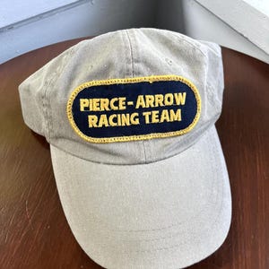 May include: A light tan baseball cap featuring a rectangular navy blue patch on the front. The patch has a yellow embroidered border and the yellow text PIERCE-ARROW RACING TEAM stitched in the center. The cap has a curved brim and stitched eyelets.