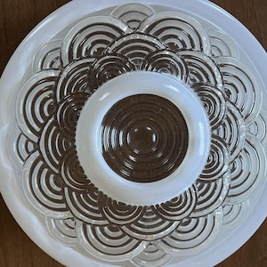 May include: A circular glass ceiling light fixture featuring a central dark brown disc surrounded by concentric rings. The glass surface is textured with a repeating pattern of overlapping semi-circular arches in clear glass, framed by a smooth white border.