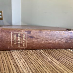 May include: A brown hardcover book titled Following the Equator by Mark Twain. The spine features a textured surface with visible wear and fraying at the edges. The title and author name are printed in white text within a rectangular border.