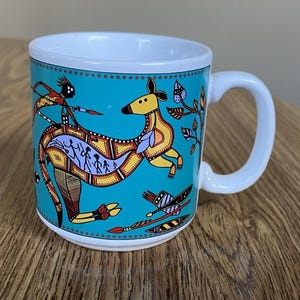 May include: A white ceramic mug featuring a vibrant turquoise background with a stylized yellow kangaroo illustration. The kangaroo's body contains intricate dot-art patterns and small human figures. Additional motifs include a hunter, foliage, and spears.