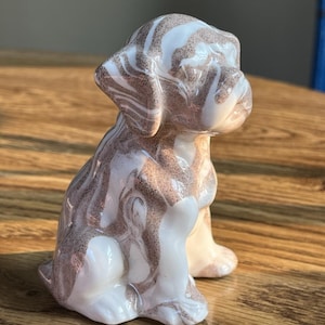May include: A small, seated puppy figurine crafted from smooth, glossy material featuring a marbled pattern of swirling white and light brown tones. The sculpture depicts a dog with floppy ears and a rounded snout, captured in a sitting position.