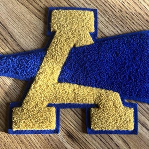 May include: A chenille patch featuring a gold letter A layered over a blue megaphone. The patch has a textured, fuzzy surface with a blue border outlining both the letter and the megaphone shape, creating a layered, collegiate-style design.